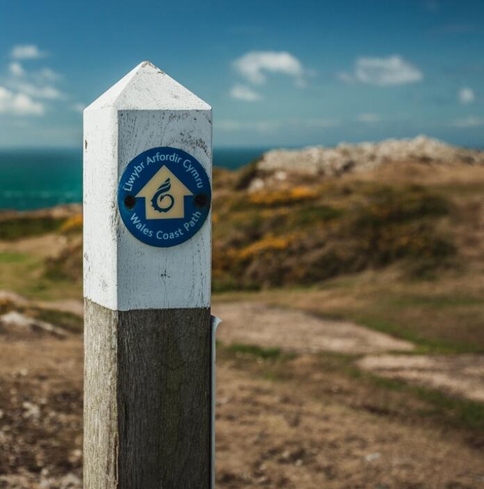 Ein hölzerner Wegweiser mit dem Abzeichen des Wales Coast Path vor dem Hintergrund des blauen Meeres und der Küsten.
