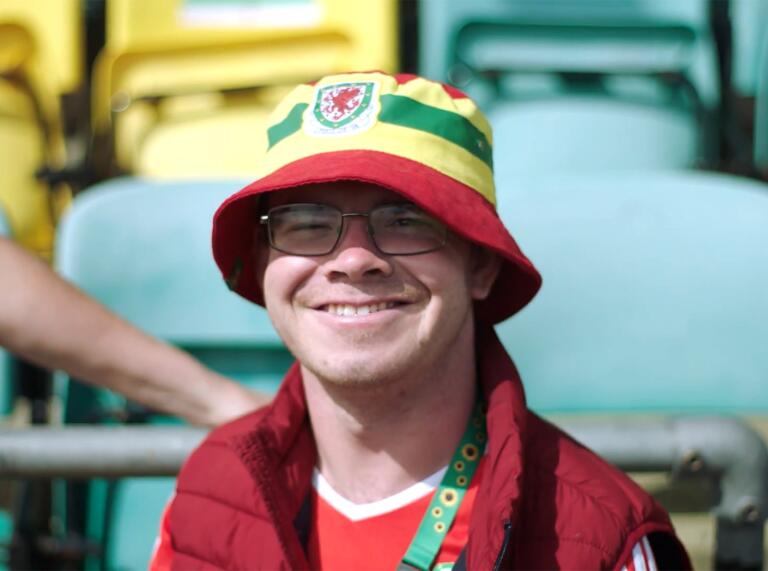 ウェールズ フットボールのトップと帽子を身にまとい、カメラに向かって微笑む男性。