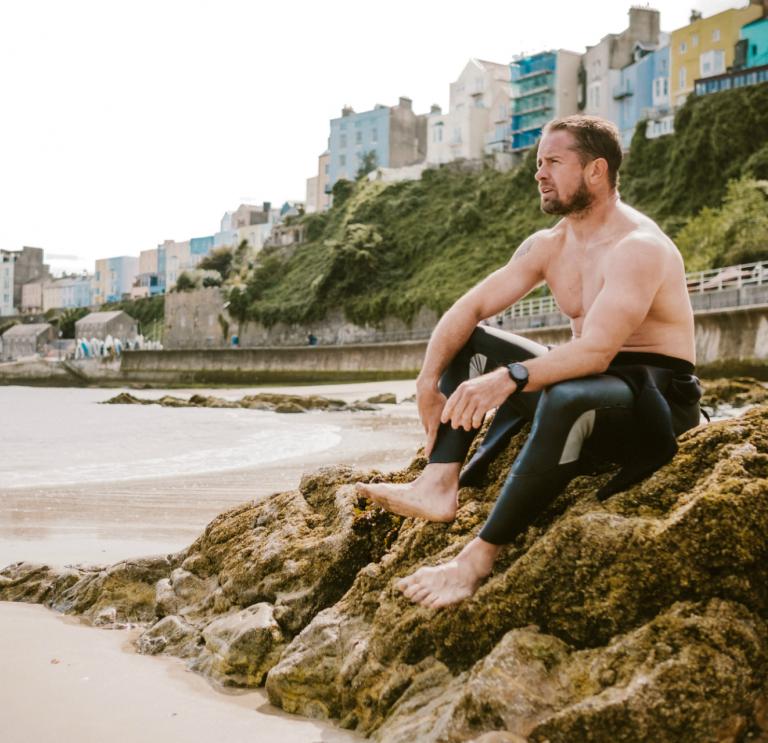 Shane Williams sitzt auf einem Felsen am North Beach in Tenby mit bunten Stadthäusern im Hintergrund.