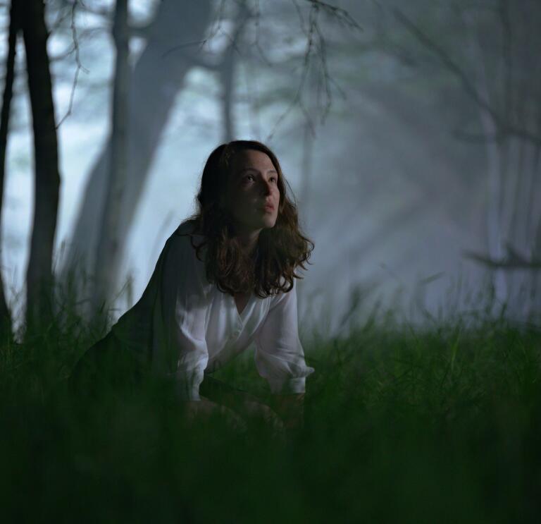 A person on their knees in the grass in a dimly lit, spooky wood at night