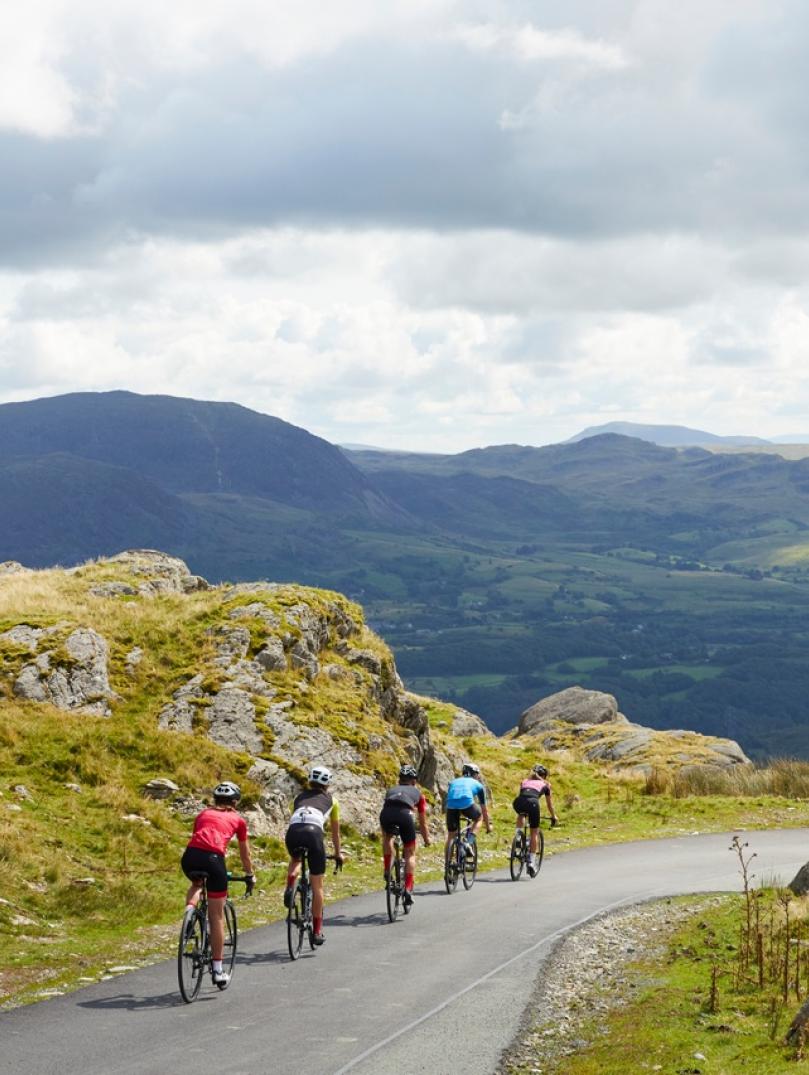Five people on road bikes cycling down a scenic road against the backdrop of mountains