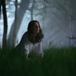A person on their knees in the grass in a dimly lit, spooky wood at night