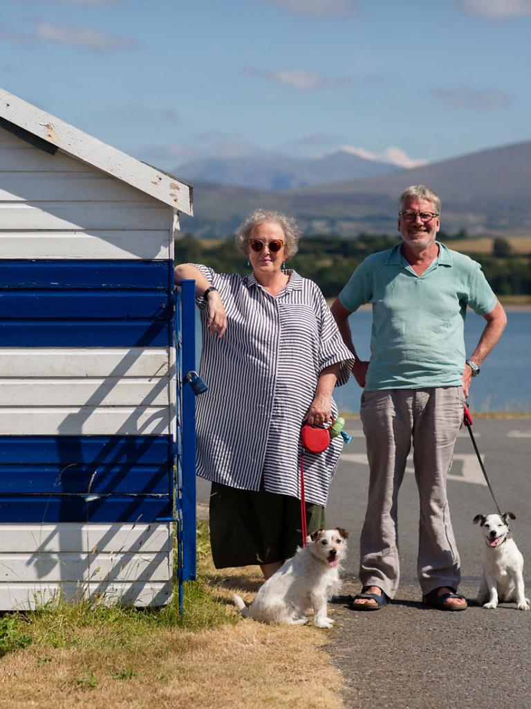 Alison and David Lea-Wilson and dogs stood next to blue and white striped shed with dogs infront of the Menai Strait