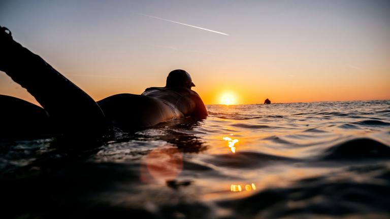 Eine Person, die auf einem Surfbrett liegt, während die Sonne untergeht und darauf wartet, eine Welle zu erwischen.