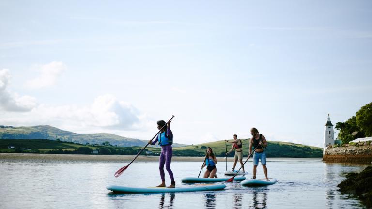 Young people standing on paddleboards in the water surrounded by soft hills and a tower. 