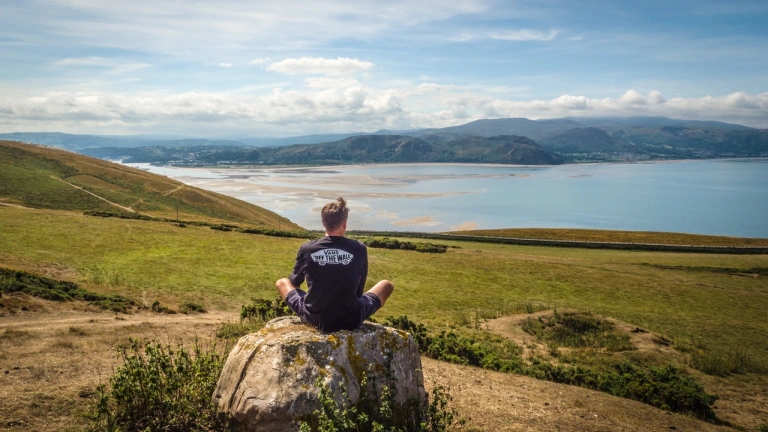 Ein Mann sitzt auf einem Felsbrocken und blickt auf Wasser und Berge.