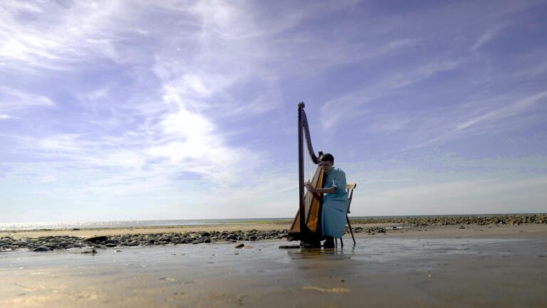 A young person wearing a blue dress playing a harp on a beach 