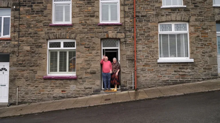 An elderly couple waving at the camera as they stand in the doorway of their home, a typical Welsh valleys house