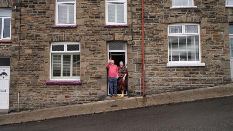 Un couple de personnes âgées, à l’entrée de leur maison typique des vallées galloises, salue la caméra.