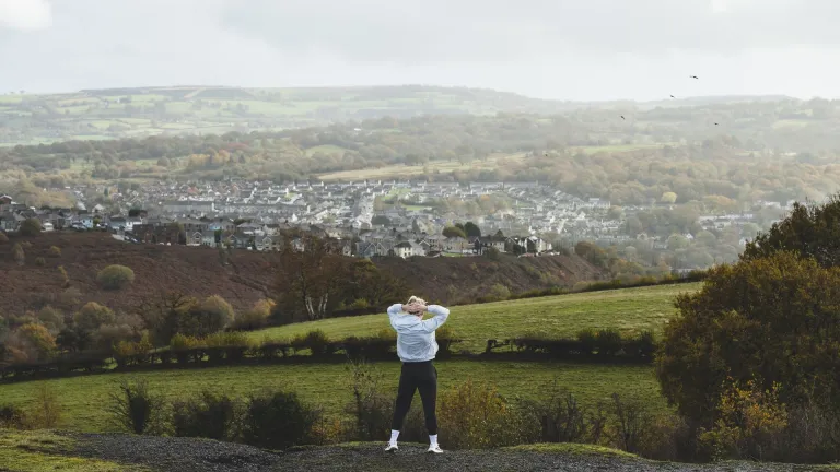 Lauren Price MBE stands on a hillside overlooking Ystrad Mynach and the surrounding valleys, with rolling green countryside stretching into the distance.