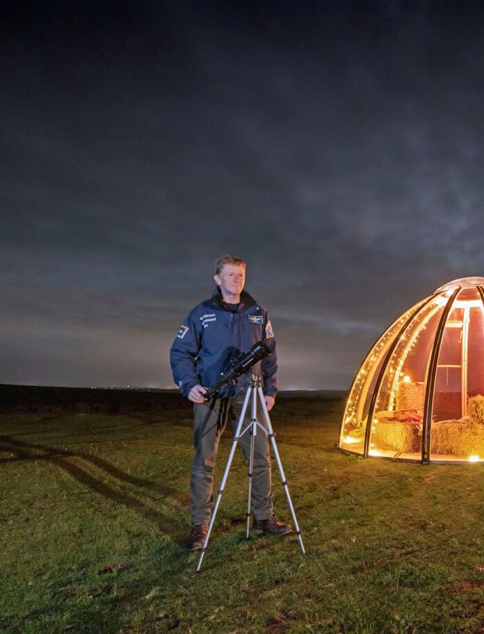 A night shot of a man with a telescope in front of a tent