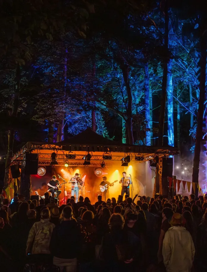 A crowded nighttime woodland concert with people standing closely together in front of a small stage, where a live band performs under warm orange lights, surrounded by tall trees illuminated in blue and green, with food stalls and festival lighting visible in the background.