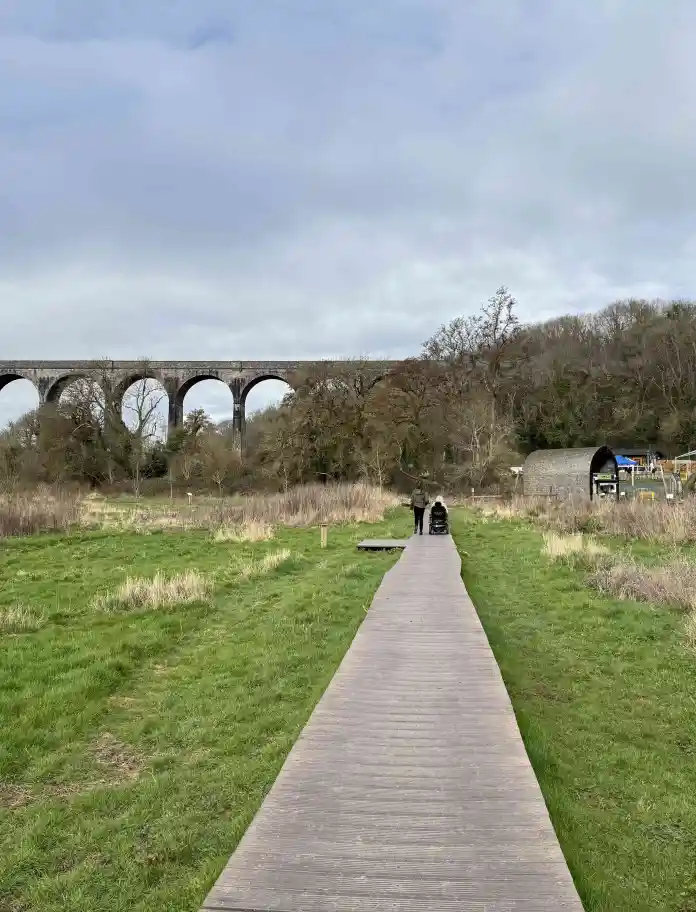 A wooden boardwalk crosses a grassy wetland towards a large stone viaduct, with two people walking and park buildings visible to the side.