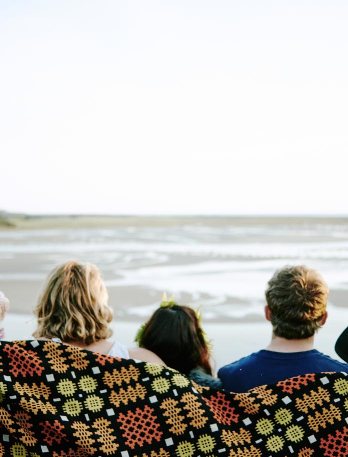 backs of heads of five people wrapped in colourful blanket with sea in background.