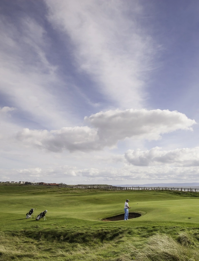 Ein Mann spielt Golf auf einem grünen weitläufigen Platz mit dem Meer im Hintergrund.