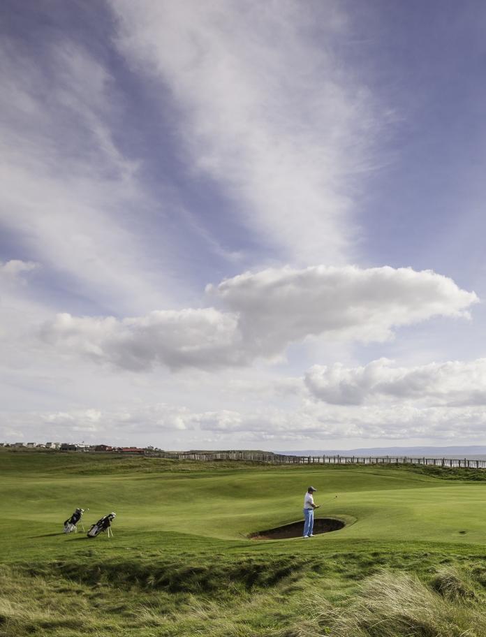 A man playing golf on a vast green course looking at the sea.