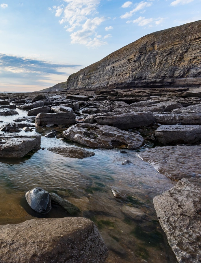 Strand in Südwales mit hohen Klippen im Hintergrund und großen Steinen im Wasser im Vordergrund.