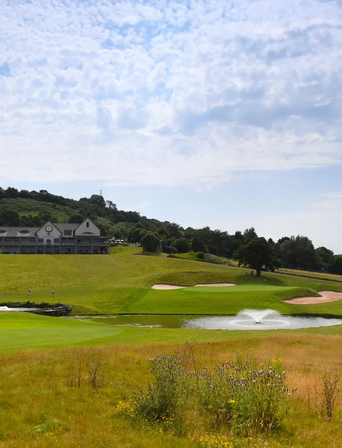 Lush green golf course featuring a water fountain, sand traps, and a distant clubhouse surrounded by rolling hills and trees.