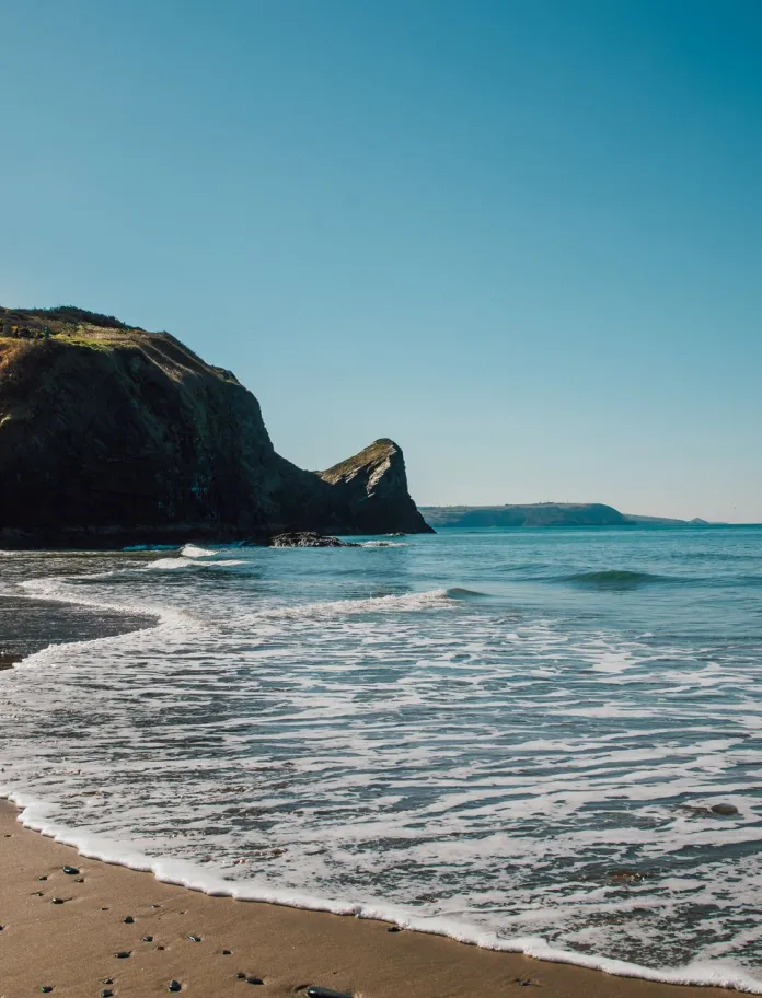 Une plage de sable où de douces vagues viennent s’échouer, un promontoire rocheux s’élevant sur la gauche, et un ciel bleu dégagé au dessus d’une mer calme.