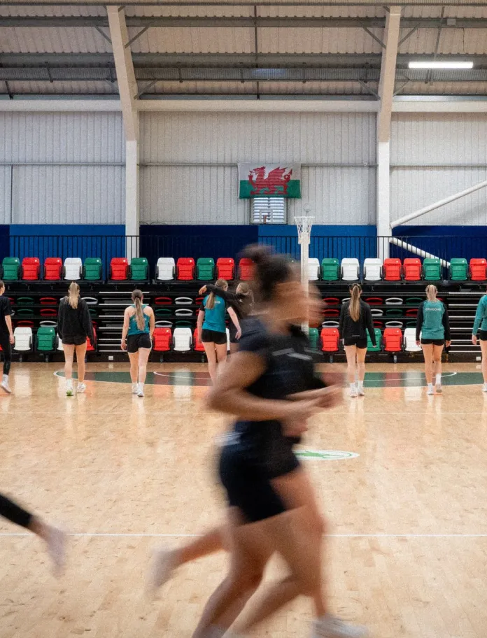 Team Wales netball players warm up on an indoor court beneath a Welsh flag ahead of training.