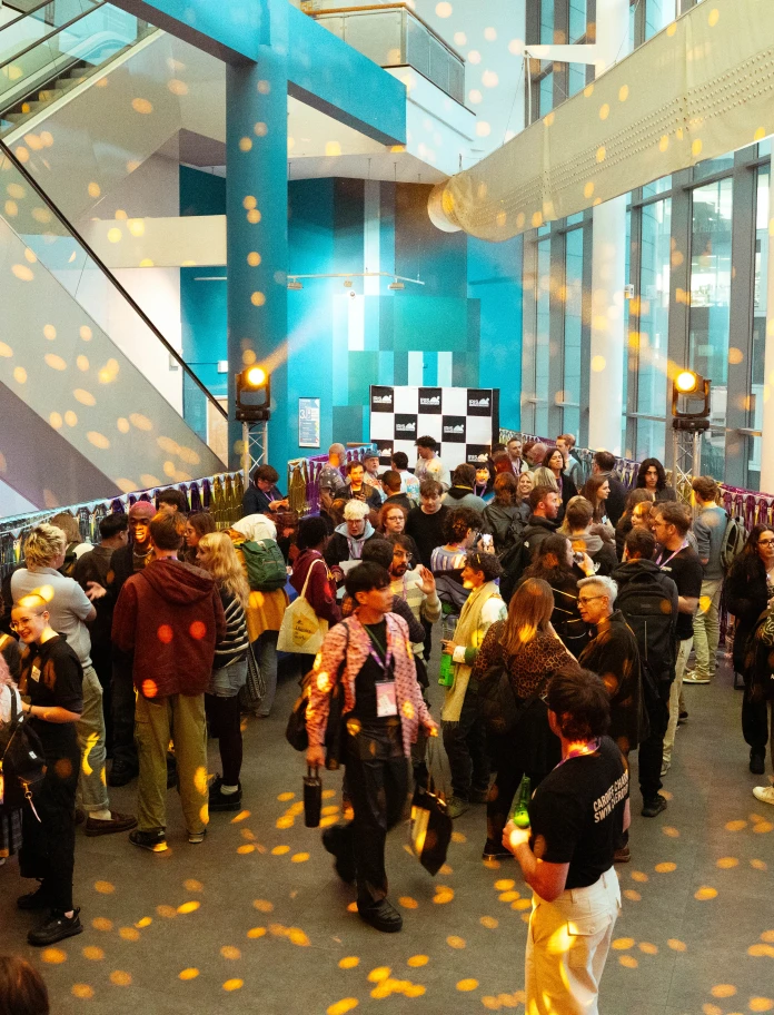 A large crowd of people mingles and socializes in a brightly lit modern building atrium decorated with hanging streamers and warm, patterned light projections. 