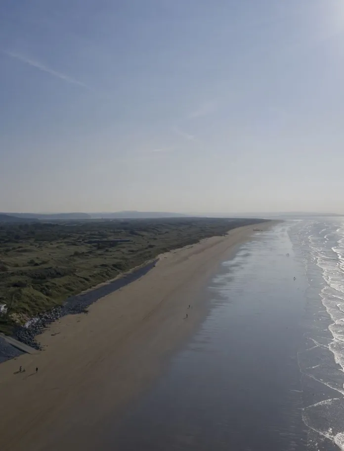 Aerial view of Pendine Sands, showing a vast sandy beach, rolling dunes and waves along the sunlit Welsh coastline. 