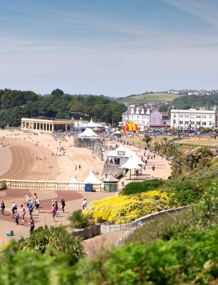 Panoramic view of Barry Island beach on a sunny day, with visitors enjoying the sandy shore, colourful fairground rides and seafront buildings. 