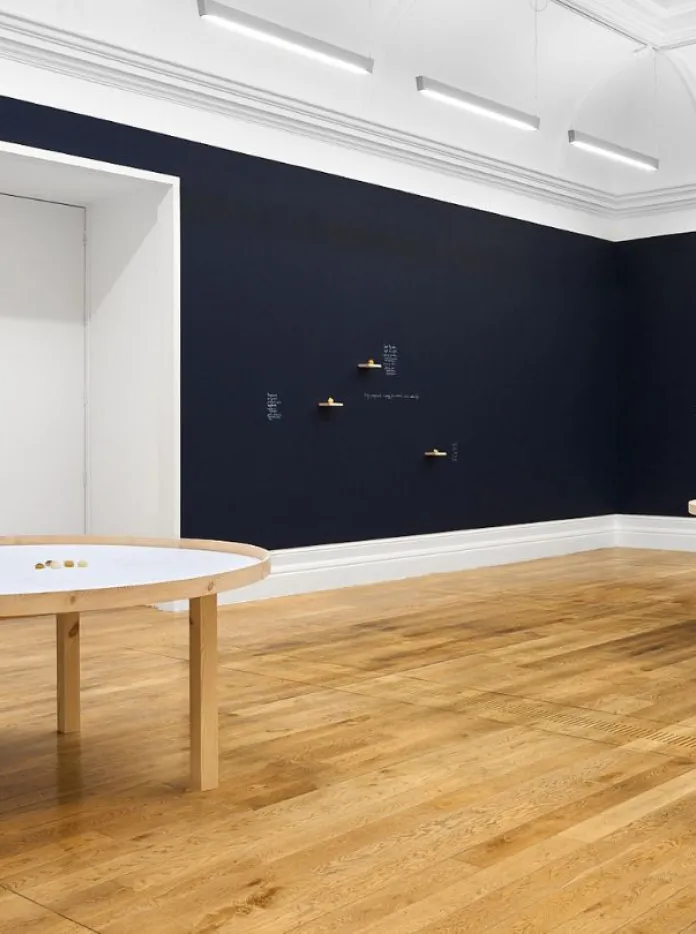 The image shows a gallery space with two round tables featuring scattered objects, and walls adorned with small sculptures and text.