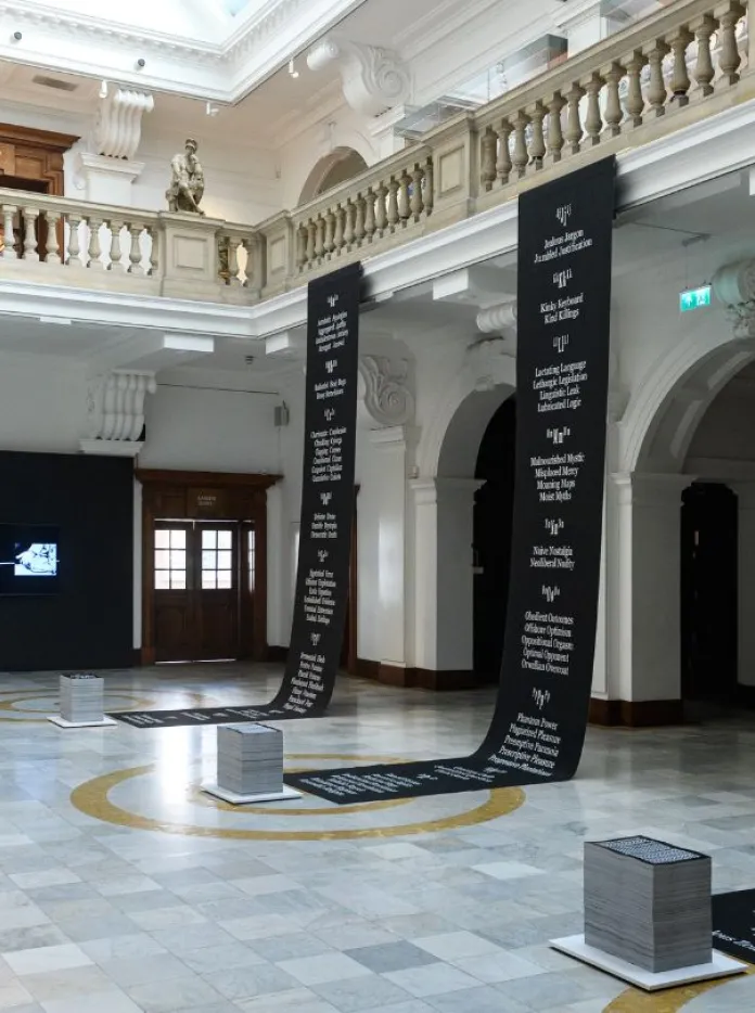 A grand hall features tall black banners printed with text, wall-mounted graphic panels, and a central video screen as part of a contemporary art installation.