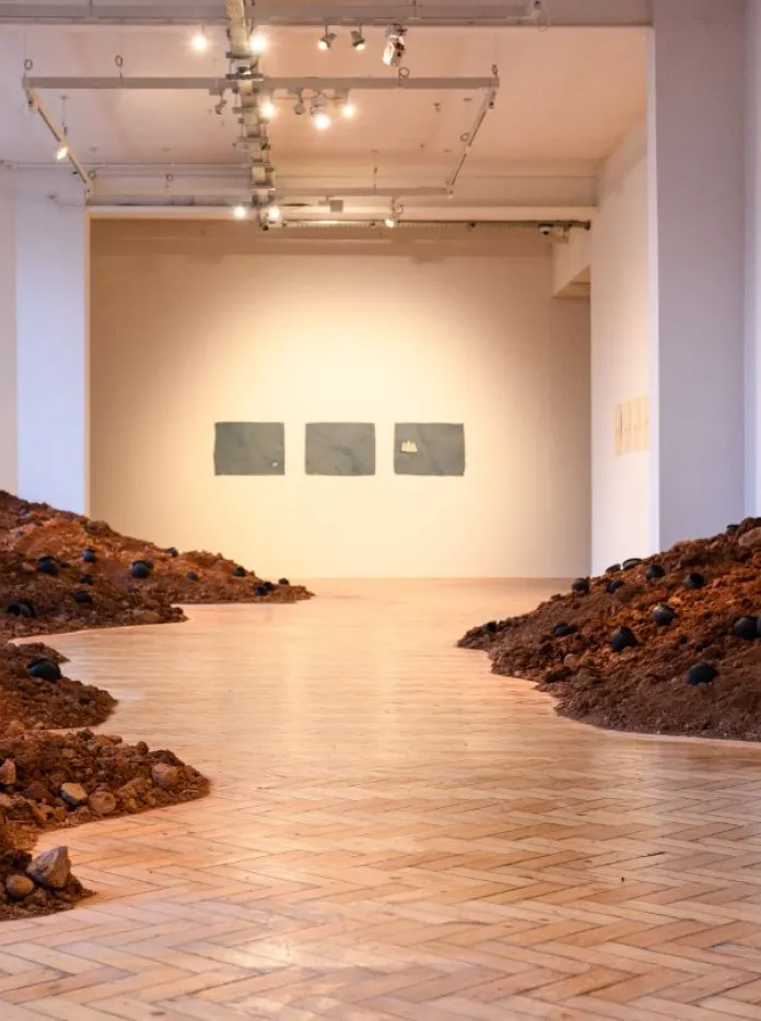 An installation of winding mounds of soil dotted with small black vessels runs through a bright gallery space toward framed works on the far wall.