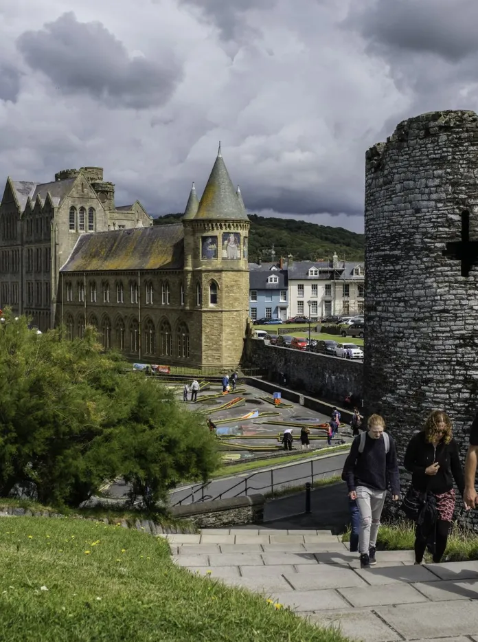 View of a historic stone castle tower and nearby university buildings in Aberystwyth, with people walking up a path in the foreground under a cloudy sky.