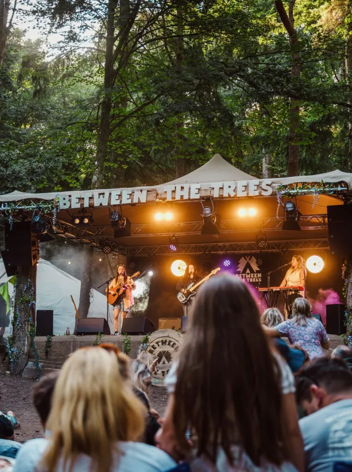 An outdoor music performance in a forest clearing, with a small stage labelled “Between the Trees” where a band plays under warm lights, while an audience sits and stands among tall trees, bunting, and festival decorations in soft daylight.