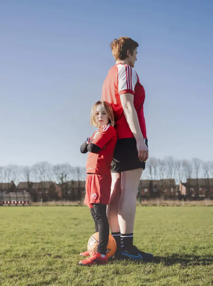Betsan Wyn Morris and her daughter Casi stand back to back on a football pitch wearing matching red football kits, with Casi resting one foot on a football and looking towards the camera beneath a clear blue sky. 