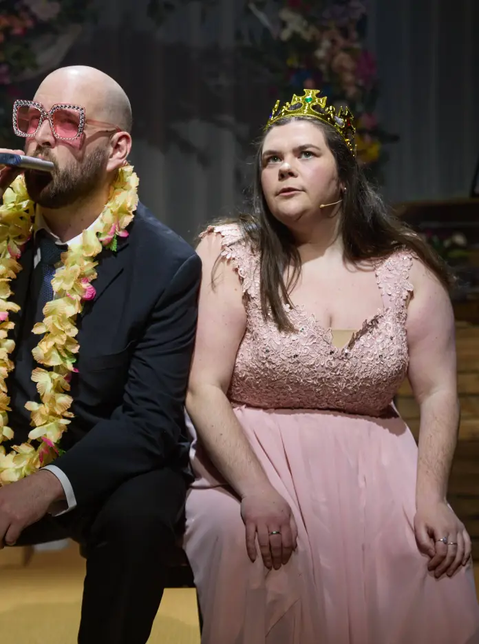 The two lead actors in the play Parti Priodas sit together on stage, with one sipping from a hipflask. 