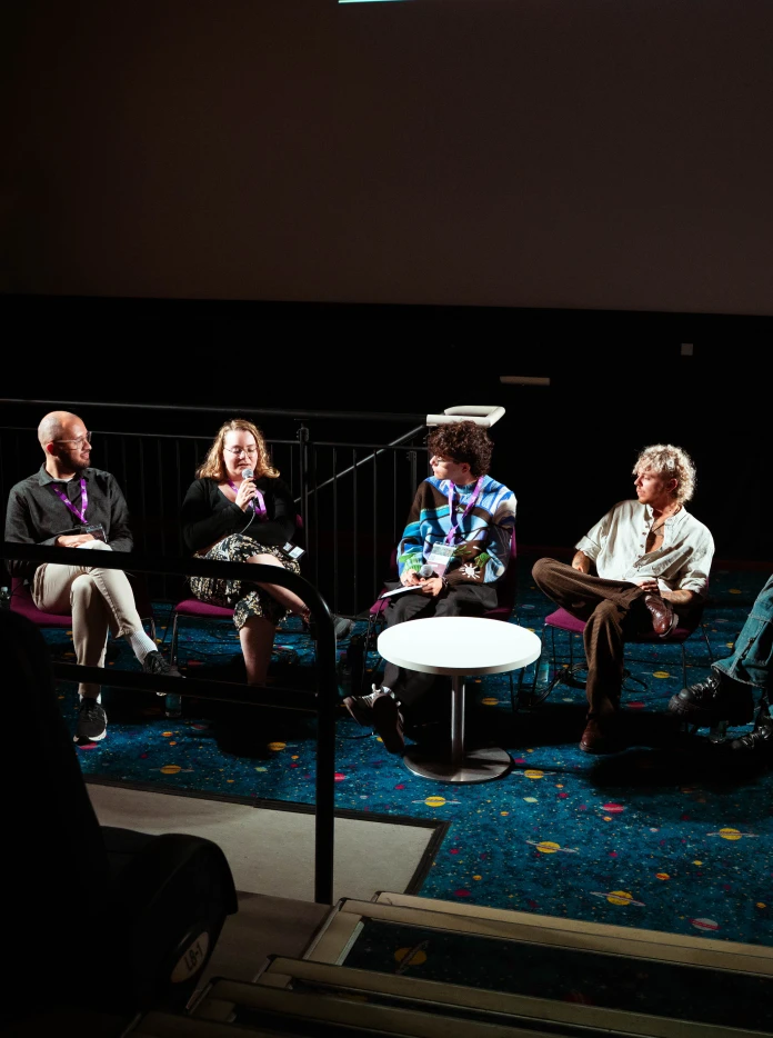 Panel discussion taking place in a cinema, with five speakers seated in front of the screen.