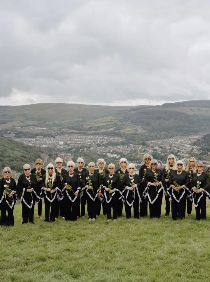 El coro femenino Dare to Sing, vestido de negro, de pie sobre un prado verde frente a los valles ondulados de Gales.