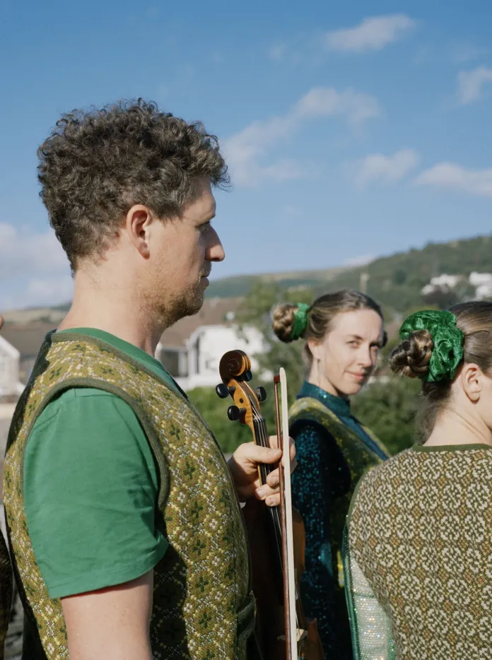 Cuatro intérpretes vestidos con trajes tradicionales galeses están de pie al aire libre bajo un cielo azul.