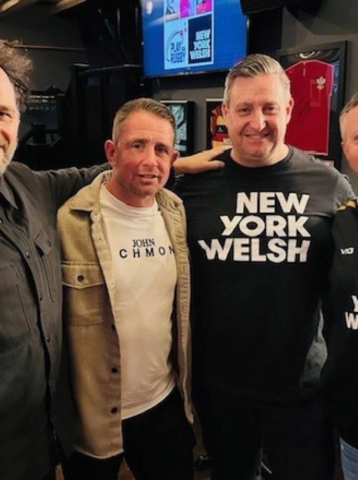 A group of four men standing together in a bar, smiling at the camera, two wearing New York Welsh shirts and one wearing a rugby style top.