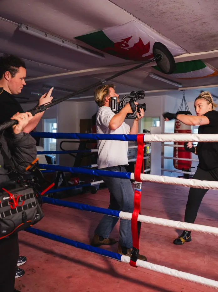 Lauren Price MBE trains in a boxing ring while a film crew records her, with a camera operator and boom microphone visible at ringside. 