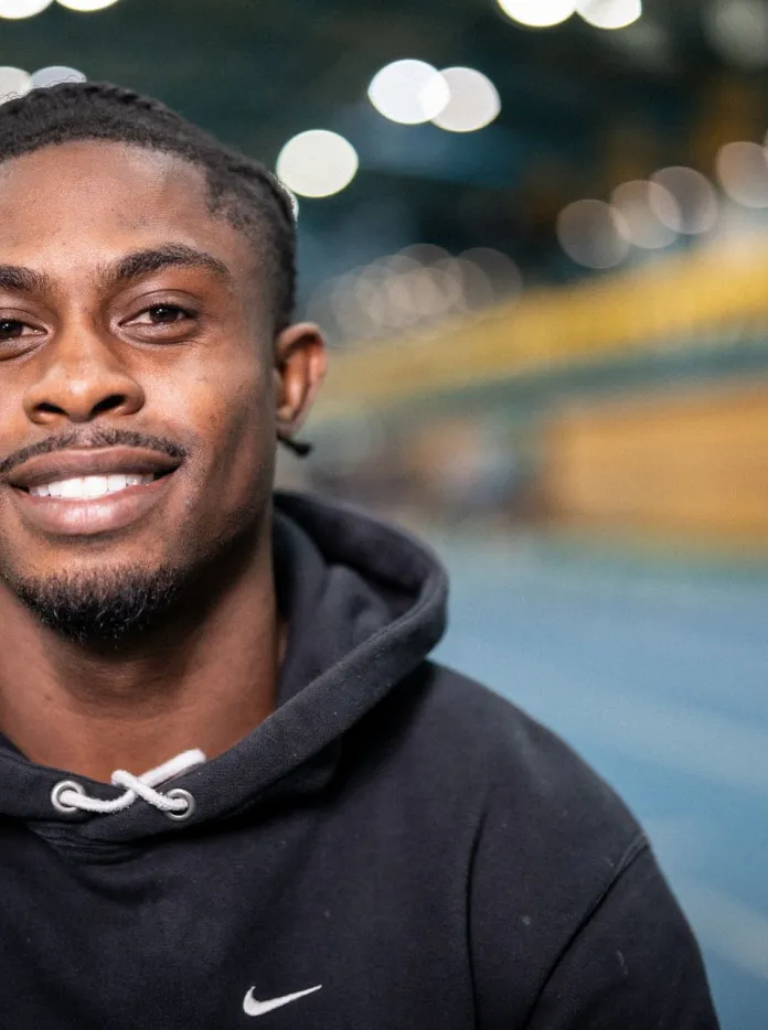 Jeremiah Azu smiling in an indoor athletics track wearing a black hoodie.