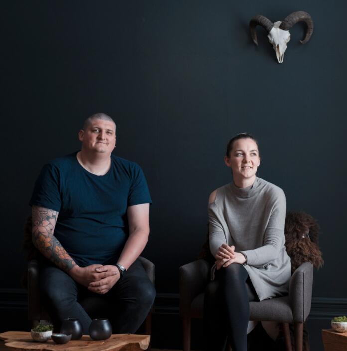 A man and a woman sitting on chairs in front of a black wall