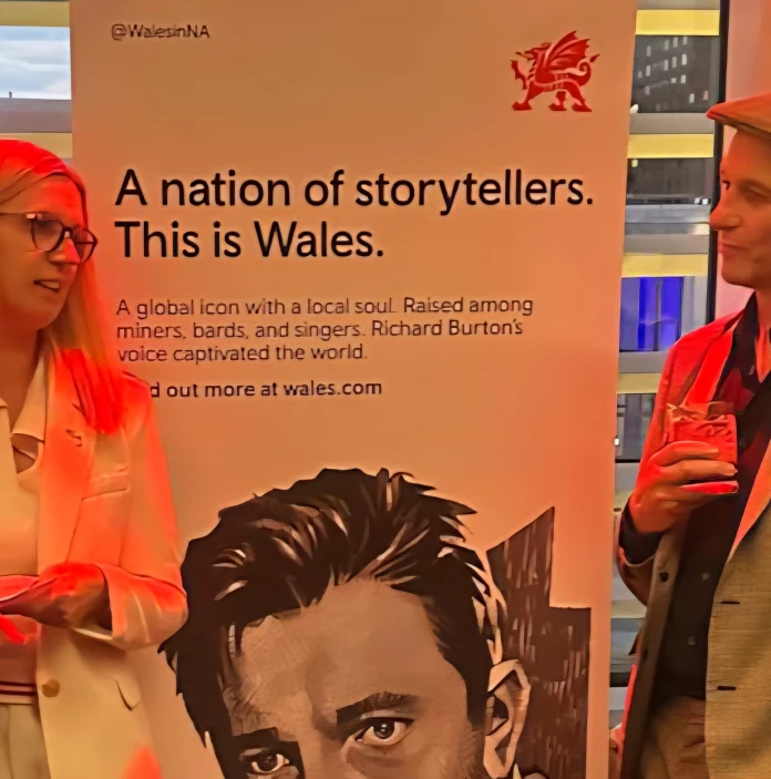 A woman and a man chat in front of a Welsh promotional banner at an indoor event.