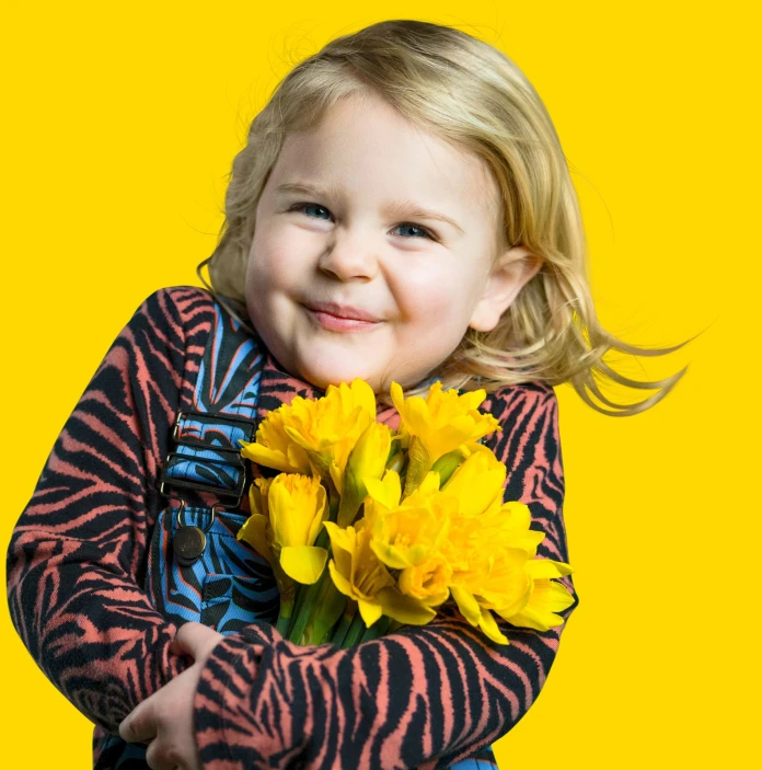 Une petite fille blonde dans une robe rose et bleue, tenant un gros bouquet de jonquilles et souriant a l’appareil