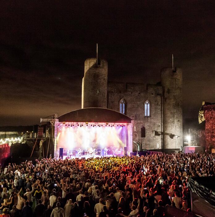 An outdoor concert stage in the grounds of a large castle with big crowds there