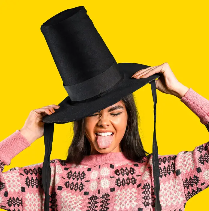Femme portant un haut chapeau gallois et un pull traditionnel gallois rose, sur un fond jaune. Elle a les yeux fermés et tire la langue
