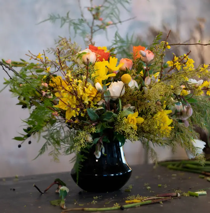 Un vase en verre vert foncé sur une table de fleuriste, rempli de jonquilles et d’autres fleurs printanières aux couleurs vives, des fleurs