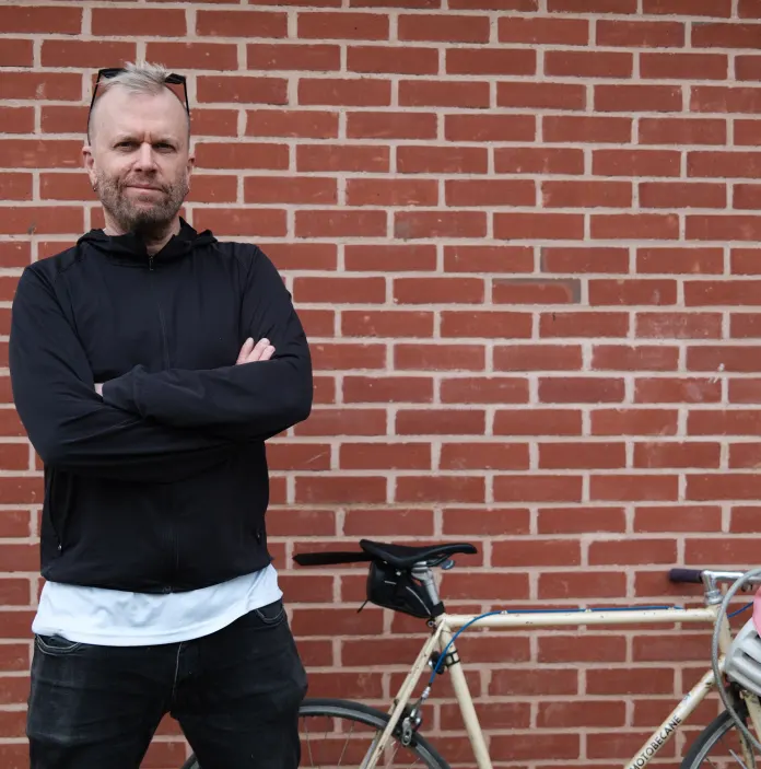Peredur ap Gwynedd standing against a brick wall with his bike