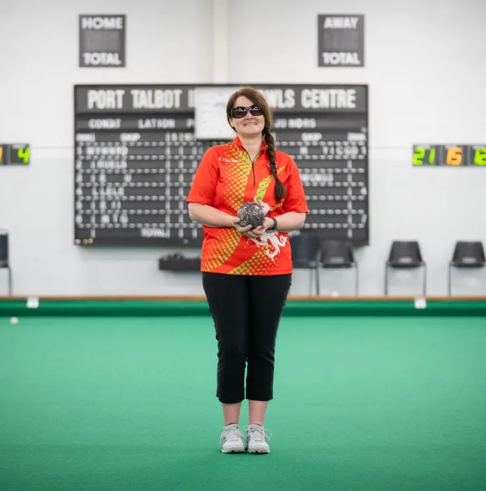Julie Thomas holding a bowls ball on an indoor bowling green in Team Wales kit.