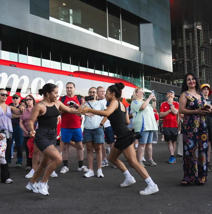 Two women dance together in front of a coach as a crowd of football fans watches.