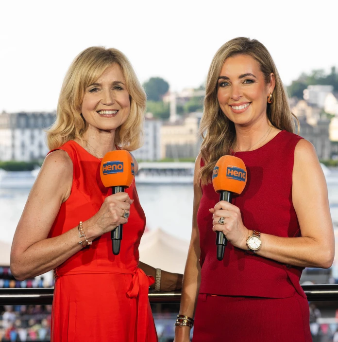 Two female presenters in red dresses smile at the camera while holding microphones beside a waterfront.